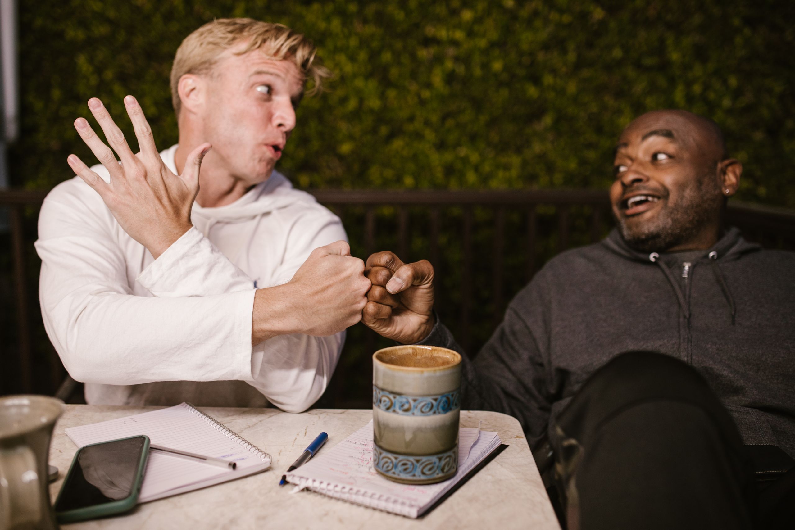 Two friends share a joyful fist bump while enjoying coffee outdoors, highlighting friendship.